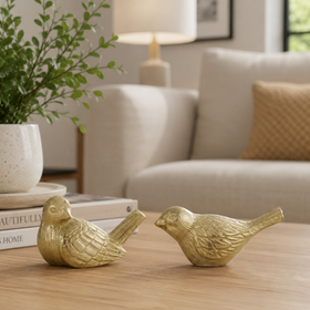 Two small gold bird figurines with textured feathers, displayed on a light wood table in a cozy living room setting next to a potted plant and books.