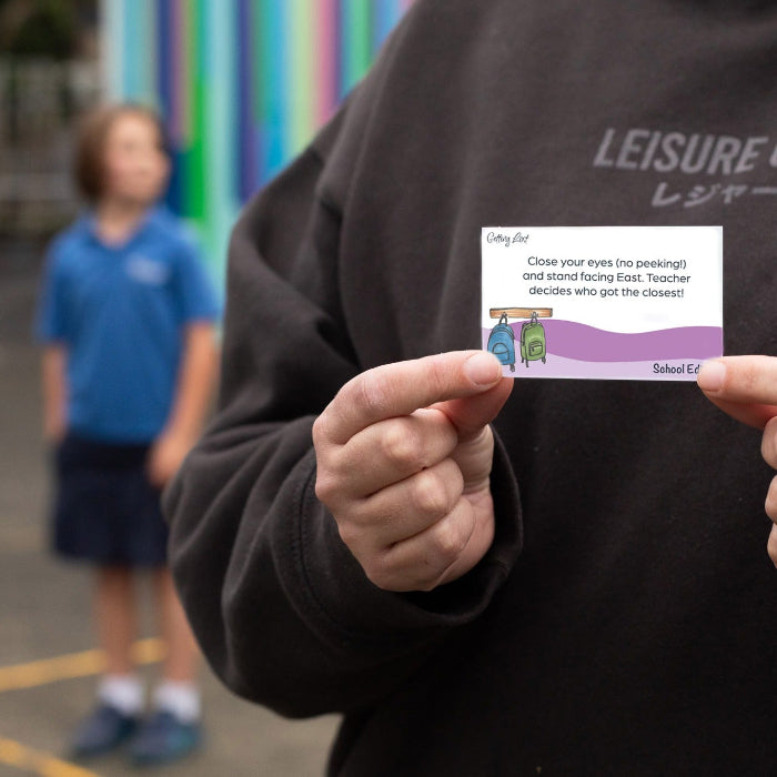 Person holding a card with text and graphics outdoors, with two children in the background.
