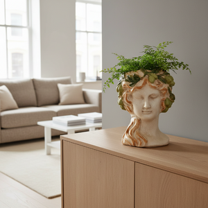 A decorative ceramic sculpture of a classical female head bust with closed eyes, wavy hair in warm tan and cream tones, and a green leafy grape crown, shown on a plain white background.