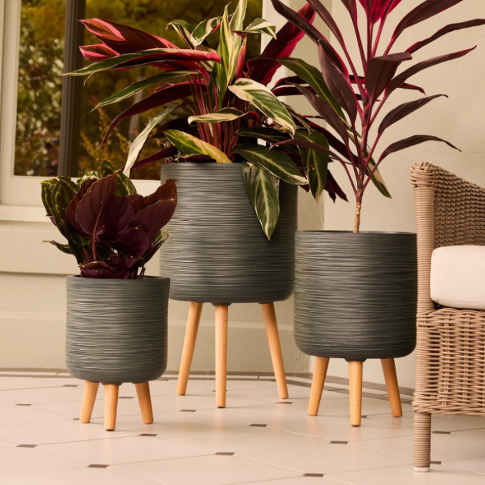 Three ribbed dark gray planters with light wood legs, each holding a different houseplant, arranged on a tiled floor next to a wicker chair.