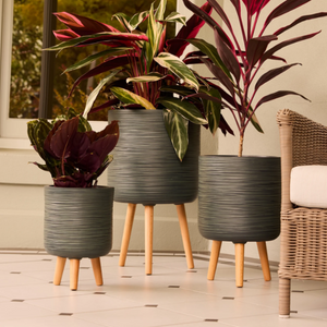 Three ribbed dark gray planters with light wood legs, each holding a different houseplant, arranged on a tiled floor next to a wicker chair.