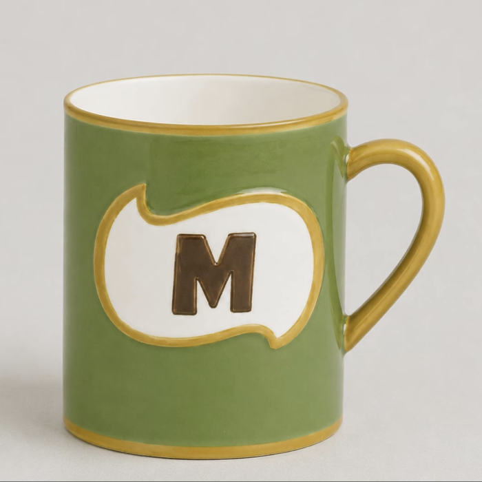 An olive green ceramic mug with a gold-trimmed handle and rim, featuring a white speech-bubble style panel on the front with a dark brown letter 'M' in the center.