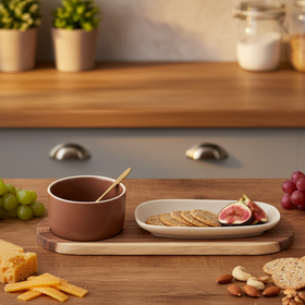 A minimalist ceramic dinnerware set on a wooden serving board, featuring a brown bowl with a gold spoon and a cream-colored oval plate, set against a plain white background.