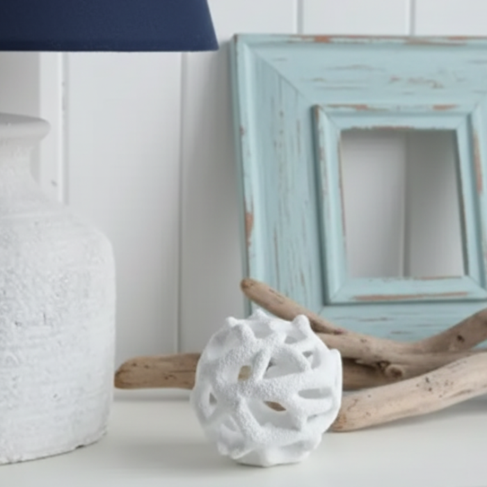 A styled interior scene with a white table lamp on the left, a white textured decorative sphere, a distressed white picture frame, and a clear glass bottle holding dried beige bunny tail grass against a white paneled wall.