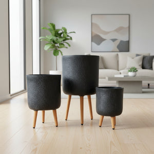 Three black textured planters with wooden legs against a brick wall.