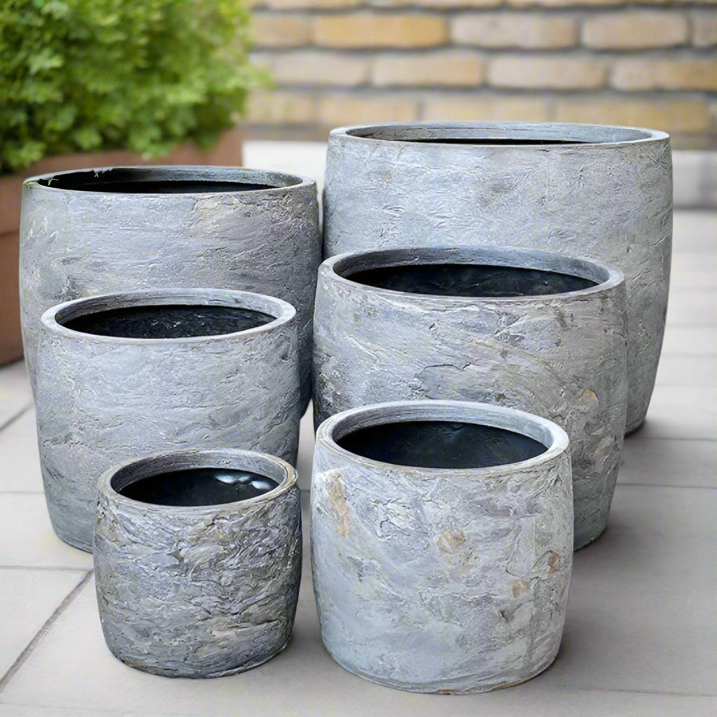 Set of gray stone-effect planters on a stone patio with plants in the background.