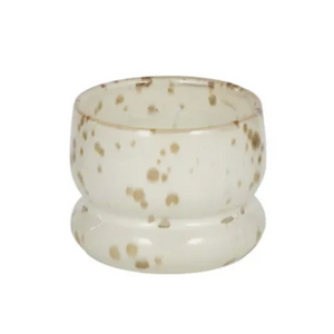 A small round glass candle holder or candle cup with a flared base, featuring a speckled pattern of light brown and beige dots on a translucent off-white glass background, isolated on a white background.