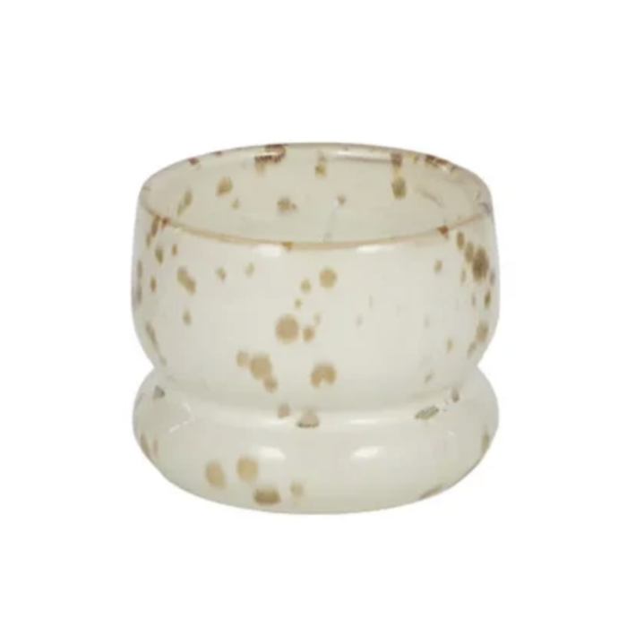 A small round glass candle holder or candle cup with a flared base, featuring a speckled pattern of light brown and beige dots on a translucent off-white glass background, isolated on a white background.