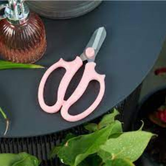 A pair of pink flower scissors resting on a dark surface with green leaves in the background.