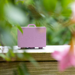 Pink suitcase with 'Gang Gang' branding on a wooden surface with blurred greenery and pink flowers in the background