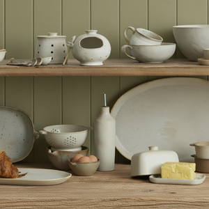 A styled collection of speckled ceramic tableware and kitchen accessories in neutral earthy tones, displayed on wooden shelves and a countertop with food props including eggs, a croissant, butter, and a plate.