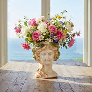 A decorative ceramic sculpture of a classical female head bust with closed eyes, wavy hair in warm tan and cream tones, and a green leafy grape crown, shown on a plain white background.