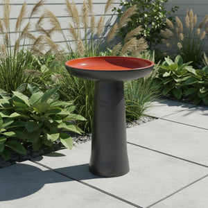 A pedestal bird bath with a dark charcoal gray base and a vibrant terracotta orange interior, shown against a plain white background with a faint line drawing of a person in the background.