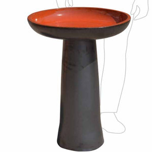 A pedestal bird bath with a dark charcoal gray base and a vibrant terracotta orange interior, shown against a plain white background with a faint line drawing of a person in the background.