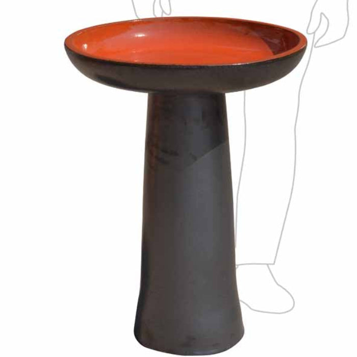 A pedestal bird bath with a dark charcoal gray base and a vibrant terracotta orange interior, shown against a plain white background with a faint line drawing of a person in the background.
