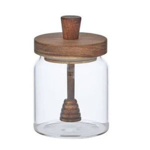 A clear glass jar with a round wooden lid and a wooden honey dipper inside, shown on a plain white background.
